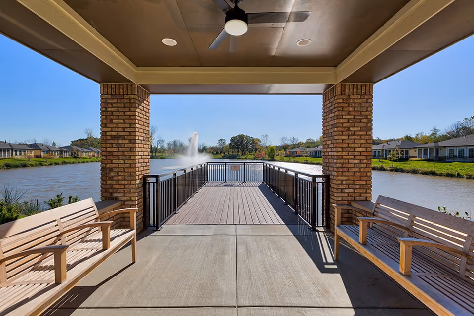 Covered patio area with two wooden benches facing a lake with a water fountain in the center. The patio has brick pillars and a ceiling fan, with a railing leading out to a wooden deck overlooking the water. Residential buildings and greenery are visible in the background under a clear blue sky.