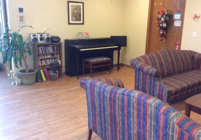 A cozy living room area with two striped upholstered sofas facing each other, a black upright piano with a cushioned bench, a small wooden shelf holding a stereo system and CDs, a potted plant, and a door decorated with a festive wreath.
