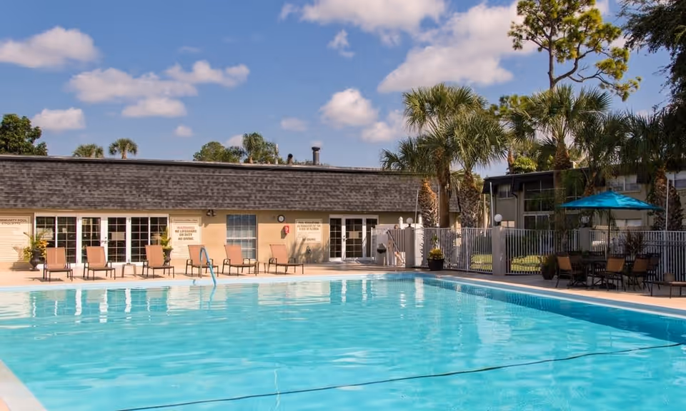 Outdoor swimming pool area at Imperial Palms facility with clear blue water, several lounge chairs lined up along the poolside, palm trees, a building with multiple doors and windows in the background, and a seating area with a blue umbrella on the right side under a partly cloudy sky.