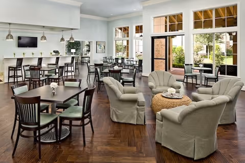 A bright and spacious dining and lounge area with multiple dark wood tables and chairs arranged for dining. There is a seating area with four cushioned armchairs around a round woven coffee table. Large windows and glass doors let in natural light and offer a view of an outdoor patio area. The room has light-colored walls, wood flooring, and pendant lights hanging over a counter with bar stools.