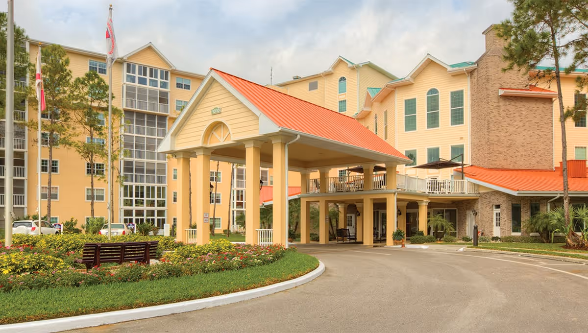 Front entrance of a multi-story senior living building with a covered porte-cochère, landscaped flowerbeds, and benches.