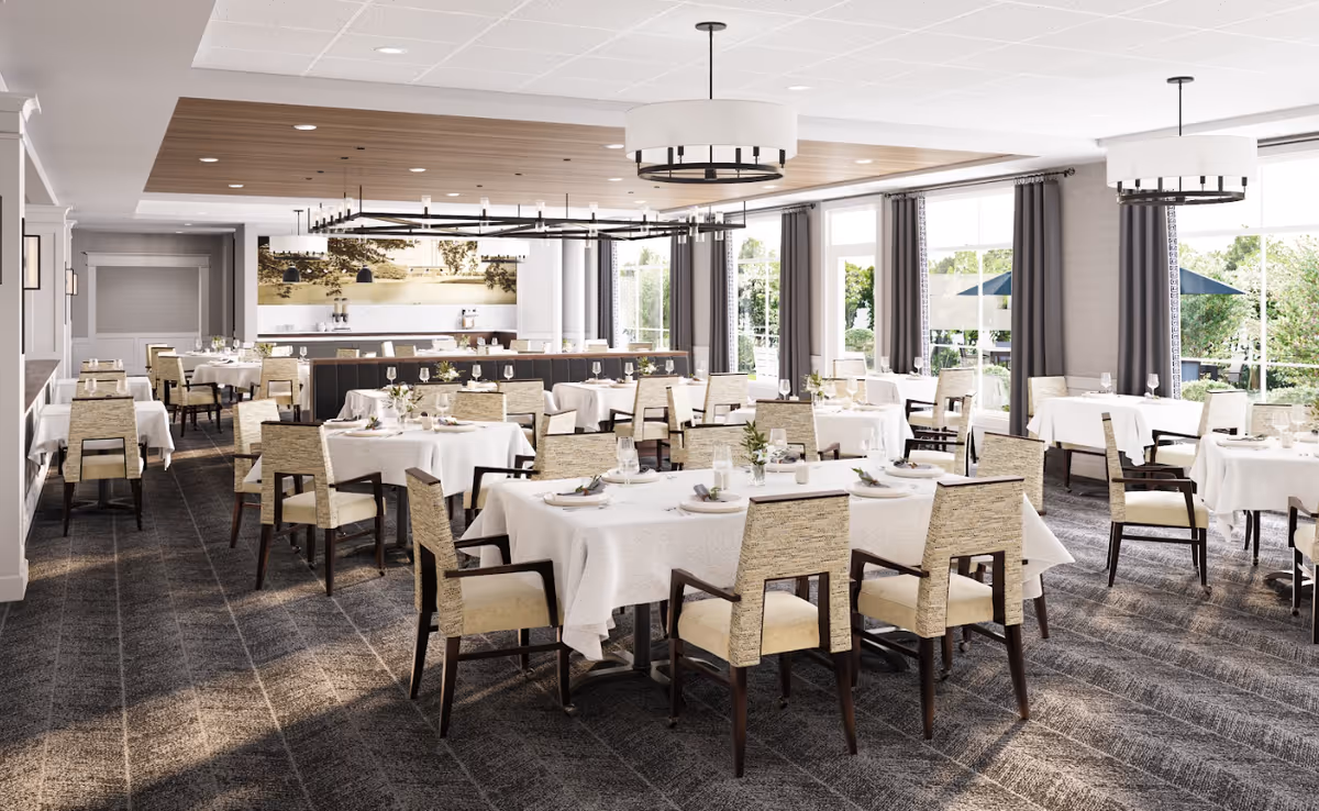 A spacious and elegant dining room with multiple tables covered in white tablecloths, each set with plates, glasses, and silverware. The room features large windows with gray curtains allowing natural light to fill the space, modern chandeliers hanging from the ceiling, and a carpeted floor with a subtle pattern. The decor is neutral and inviting, suitable for a senior living facility.