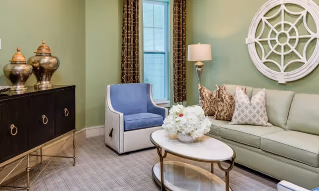 A cozy living room with a light green sofa adorned with patterned pillows, a blue and white armchair, a white oval coffee table with a flower arrangement, a floor lamp, and a decorative round wall mirror. There is a window with patterned curtains and a dark wooden cabinet with decorative vases on top.