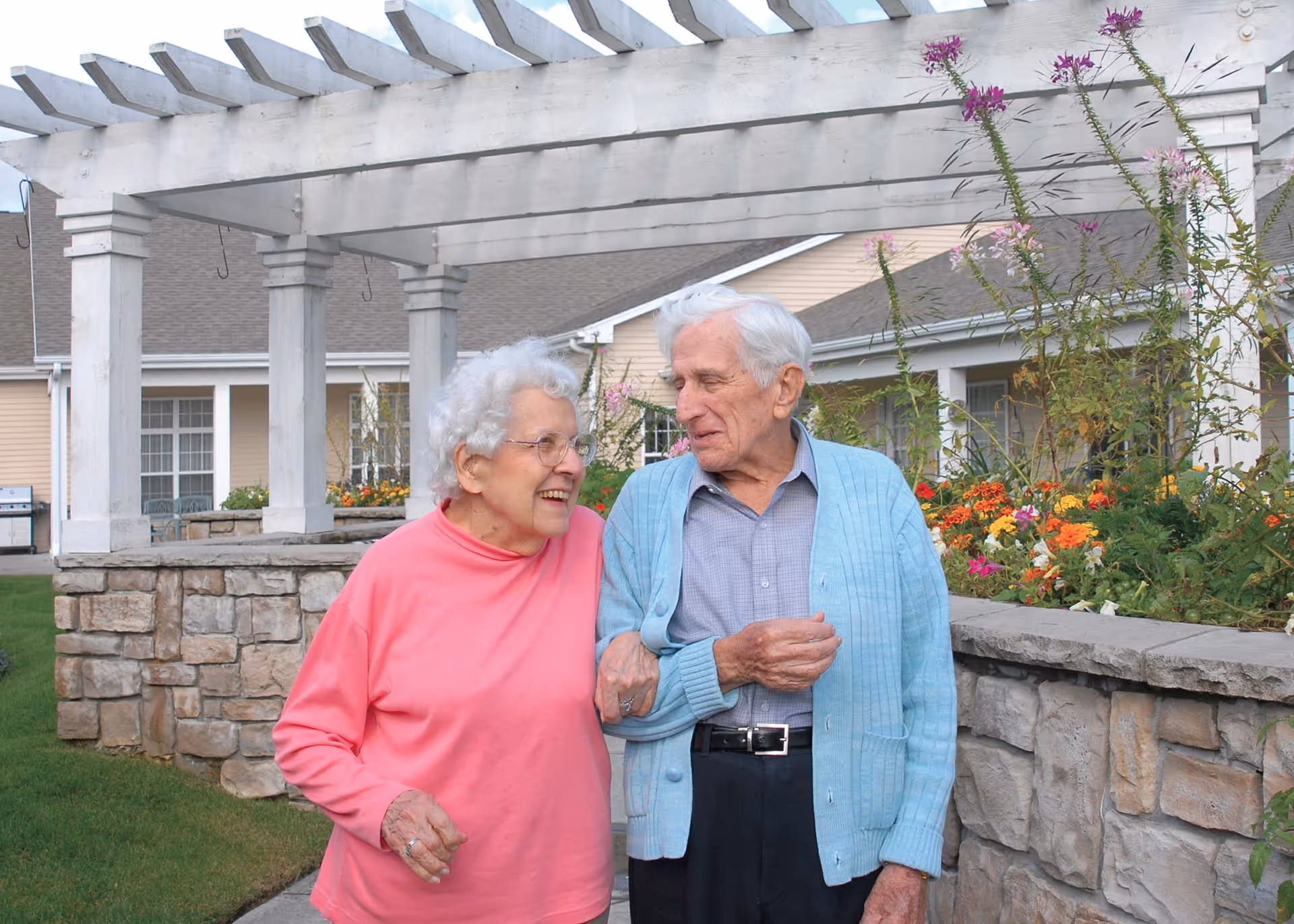 An elderly couple walking arm in arm outdoors near a stone wall and flower garden, with a white pergola and residential buildings in the background. The woman is wearing a pink sweater and glasses, and the man is wearing a light blue cardigan over a gray shirt.