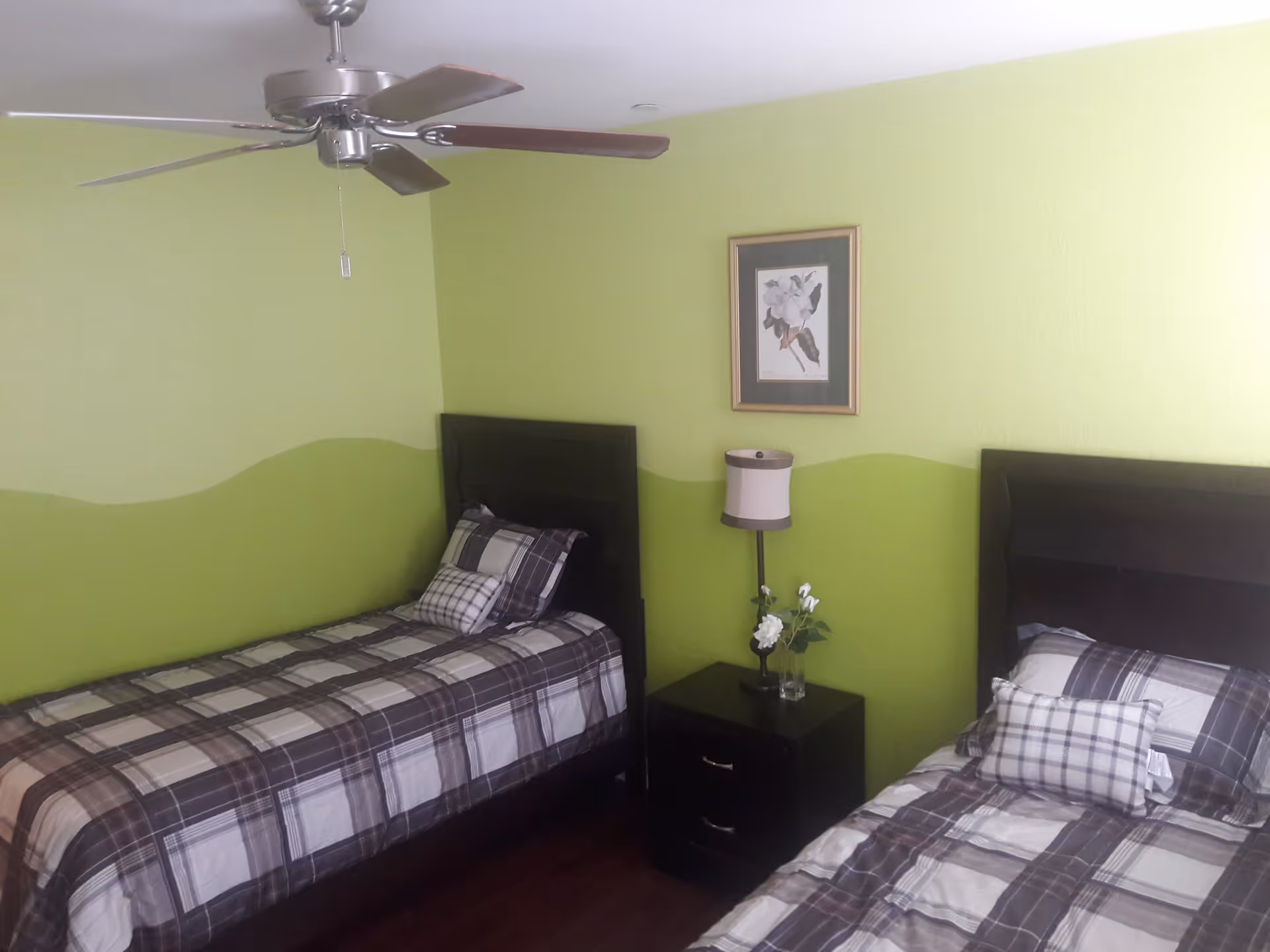 A bedroom with two single beds featuring dark wooden headboards and plaid bedding. Between the beds is a dark wooden nightstand with a small lamp and a vase with white flowers. The walls are painted light green with a darker green wavy design along the middle. A ceiling fan with wooden blades is mounted on the ceiling, and a framed floral picture hangs on the wall above the nightstand.