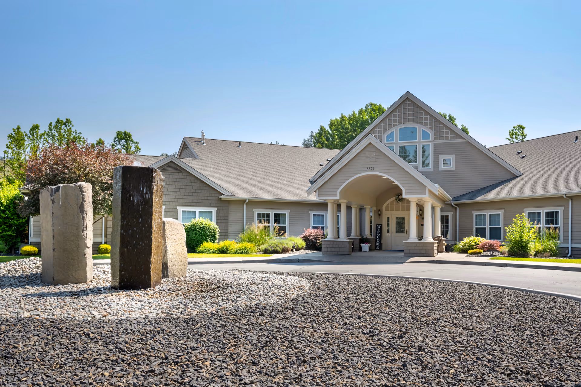 Front exterior view of Brookdale Nine Mile facility showing a beige building with a peaked roof, a covered entrance with columns, landscaped greenery, and a stone sculpture in the foreground under a clear blue sky.