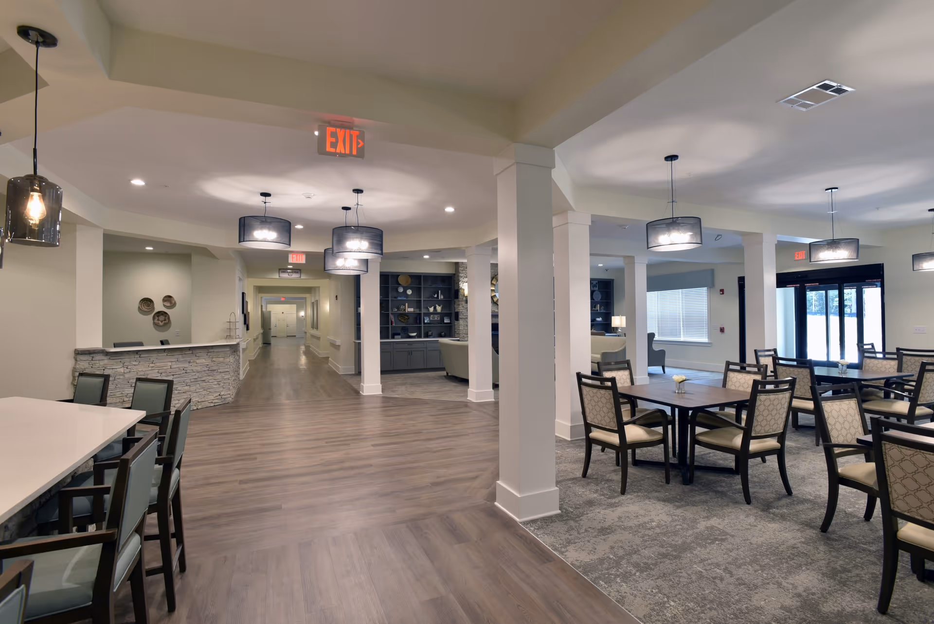 Interior view of a senior living facility showing a spacious common area with multiple dining tables and chairs on the right, a reception desk with stone facade on the left, and a hallway leading further into the building. The area is well-lit with modern pendant lights and ceiling lights, and large windows allow natural light to enter.