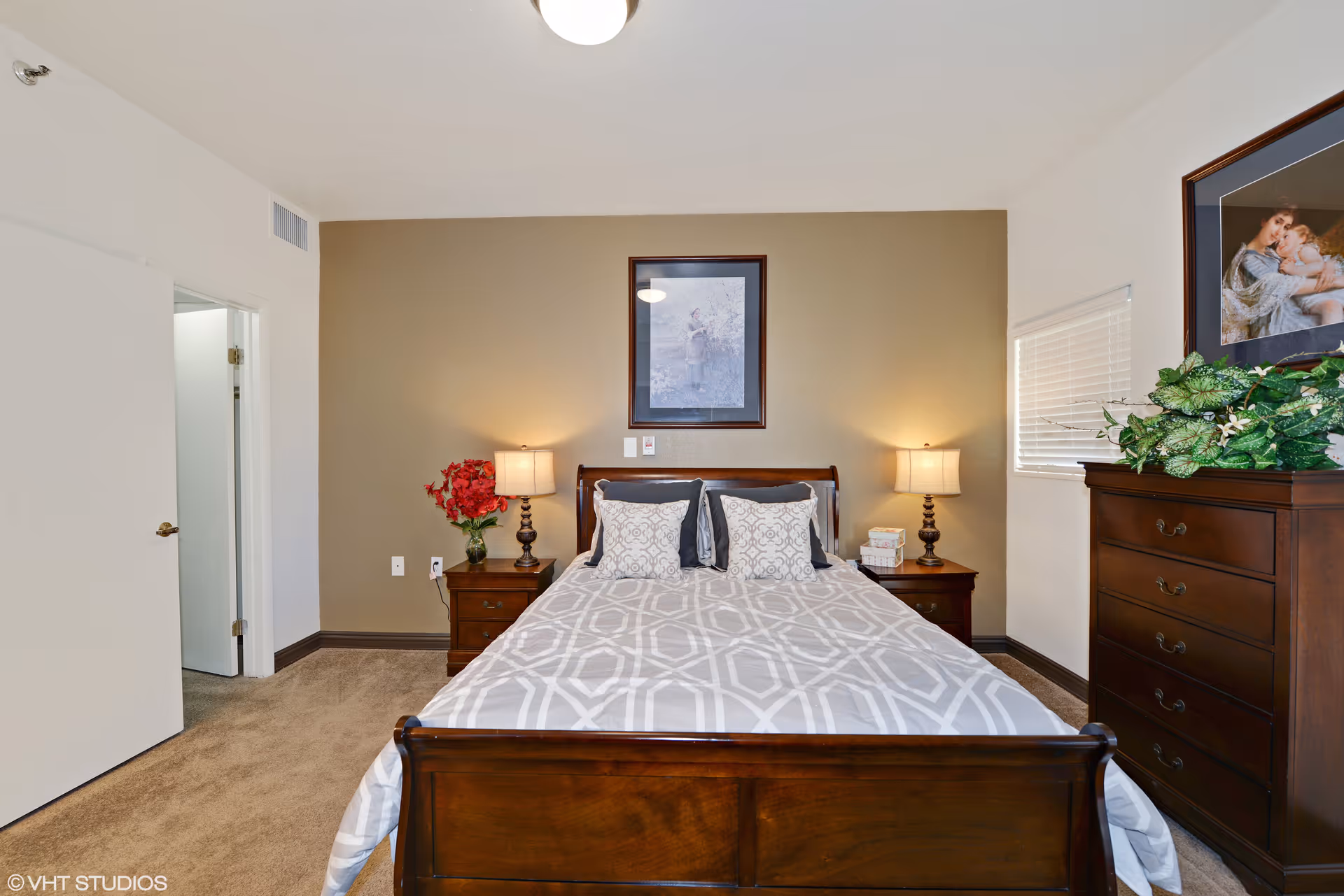 A neatly arranged bedroom with a wooden bed frame and a bed made with a gray and white patterned comforter and pillows. There are two wooden nightstands on either side of the bed, each with a lamp. A vase with red flowers is on the left nightstand, and a stack of books is on the right. A framed picture hangs on the beige accent wall behind the bed. To the right is a wooden dresser with a green leafy plant on top and another framed picture above it. The room has beige carpet and white walls, with a partially open door leading to another room.