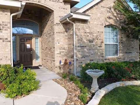 Brick senior living facility entrance with arched doorway, landscaping, and a birdbath.
