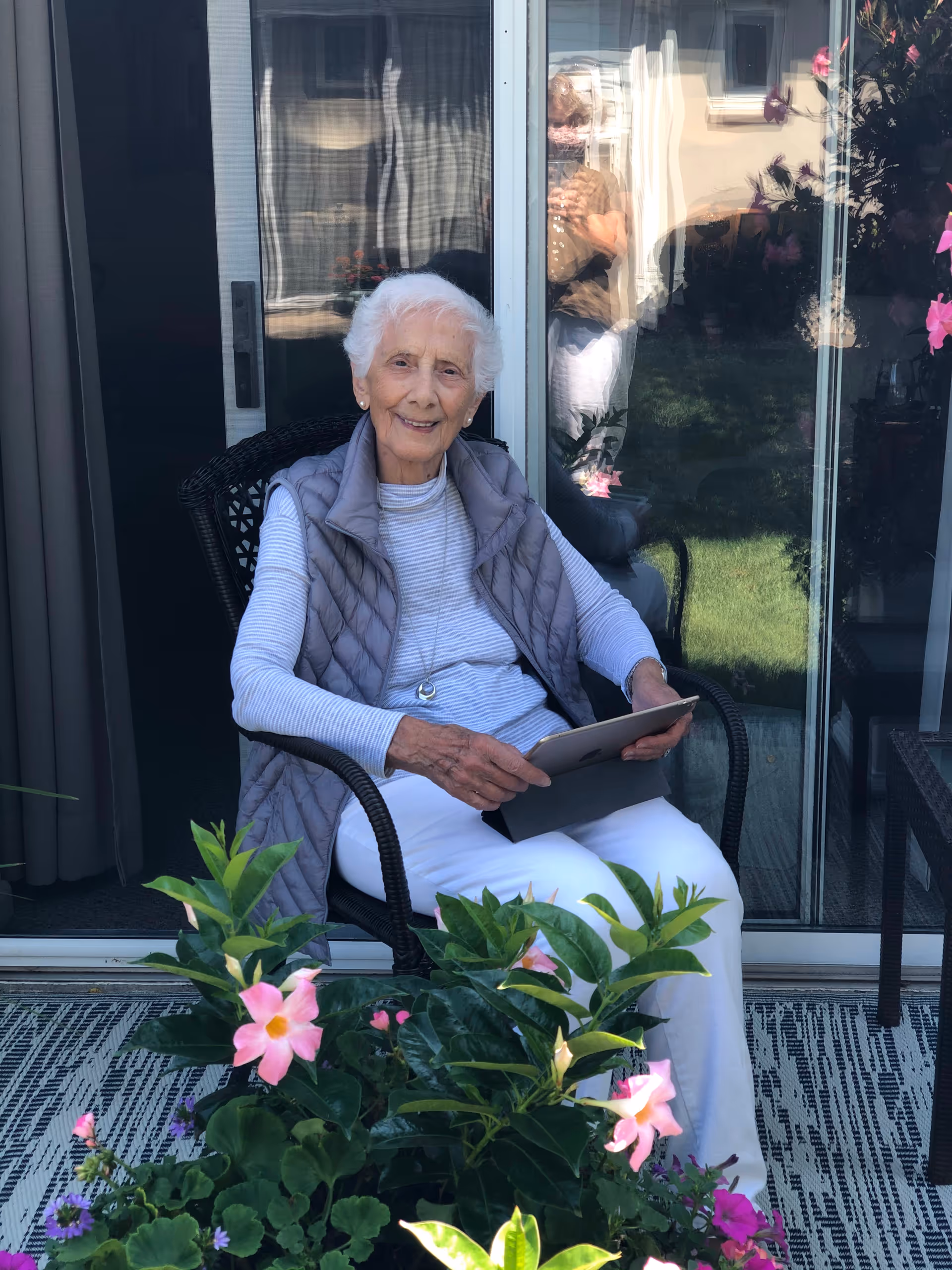 An elderly woman with white hair is sitting on a black wicker chair on a patio. She is wearing a gray quilted vest over a striped long-sleeve shirt and white pants, holding a tablet. In front of her are green plants with pink flowers. Behind her is a glass sliding door reflecting another person taking the photo and some outdoor greenery.