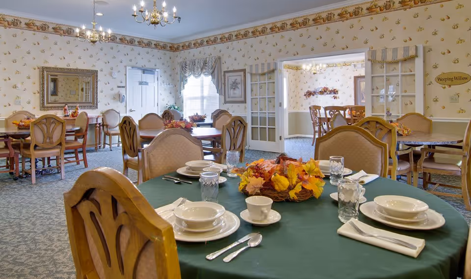 A senior living facility dining room with multiple wooden tables and chairs. One table in the foreground is set with white plates, bowls, cups, silverware, and glasses, and has a centerpiece of autumn leaves. The room has patterned wallpaper, carpeted floors, chandeliers, and a window with curtains. A sign on the wall reads 'Weeping Willow'.