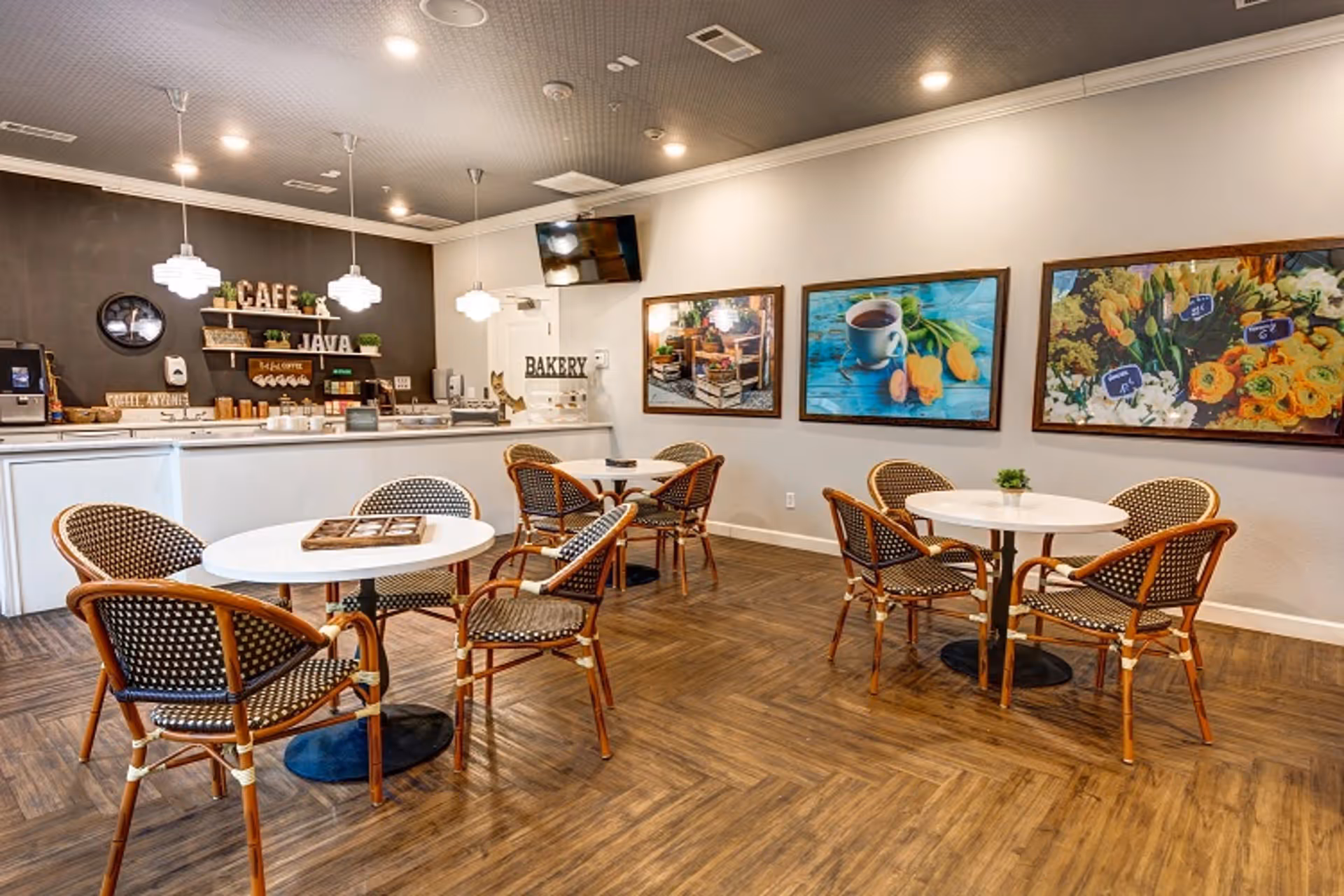 Bright cafe-style dining area with round tables and wicker chairs facing a counter labeled 'Cafe' and 'Bakery'.