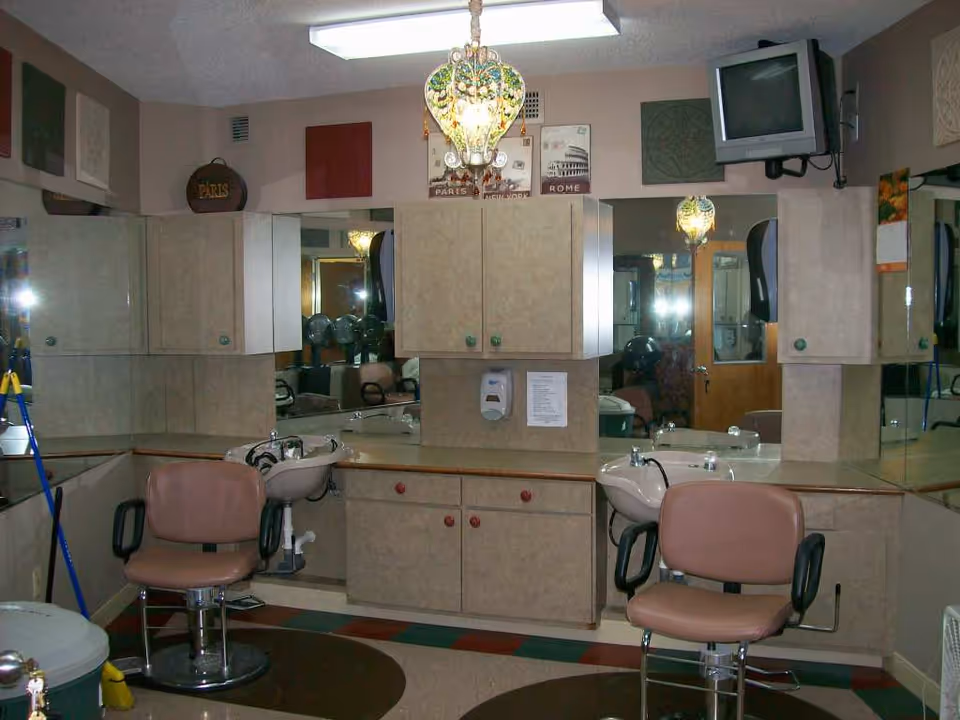 Interior view of a salon area with two salon chairs in front of sinks for hair washing, beige cabinets mounted on the walls, large mirrors, a small wall-mounted TV in the corner, and a decorative hanging light fixture.