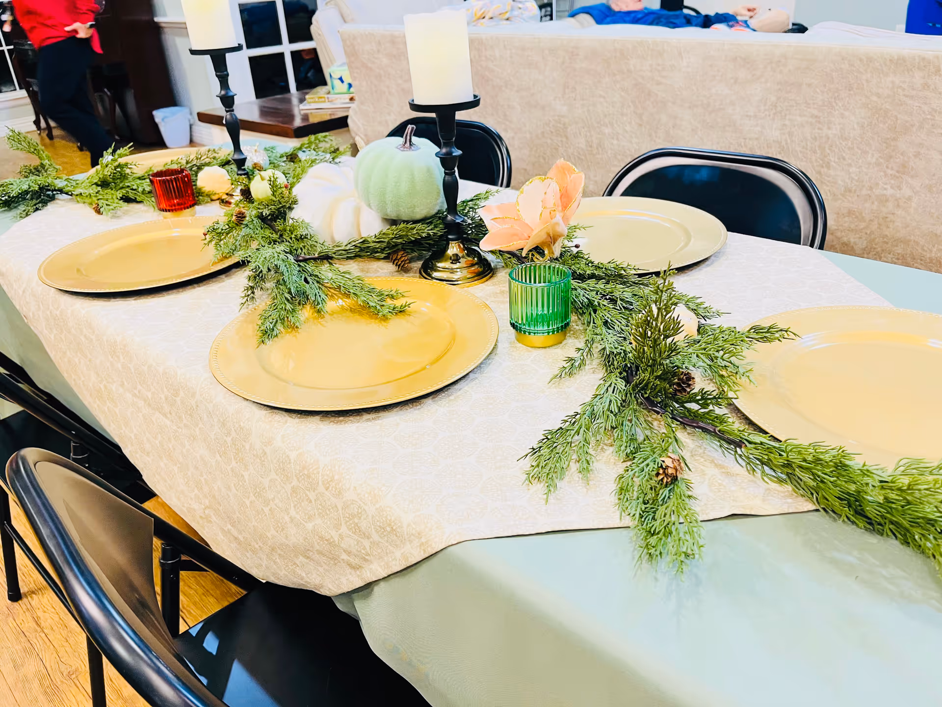 Decoratively set dining table with gold plates, greenery garland, candles and small pumpkins in a communal room.