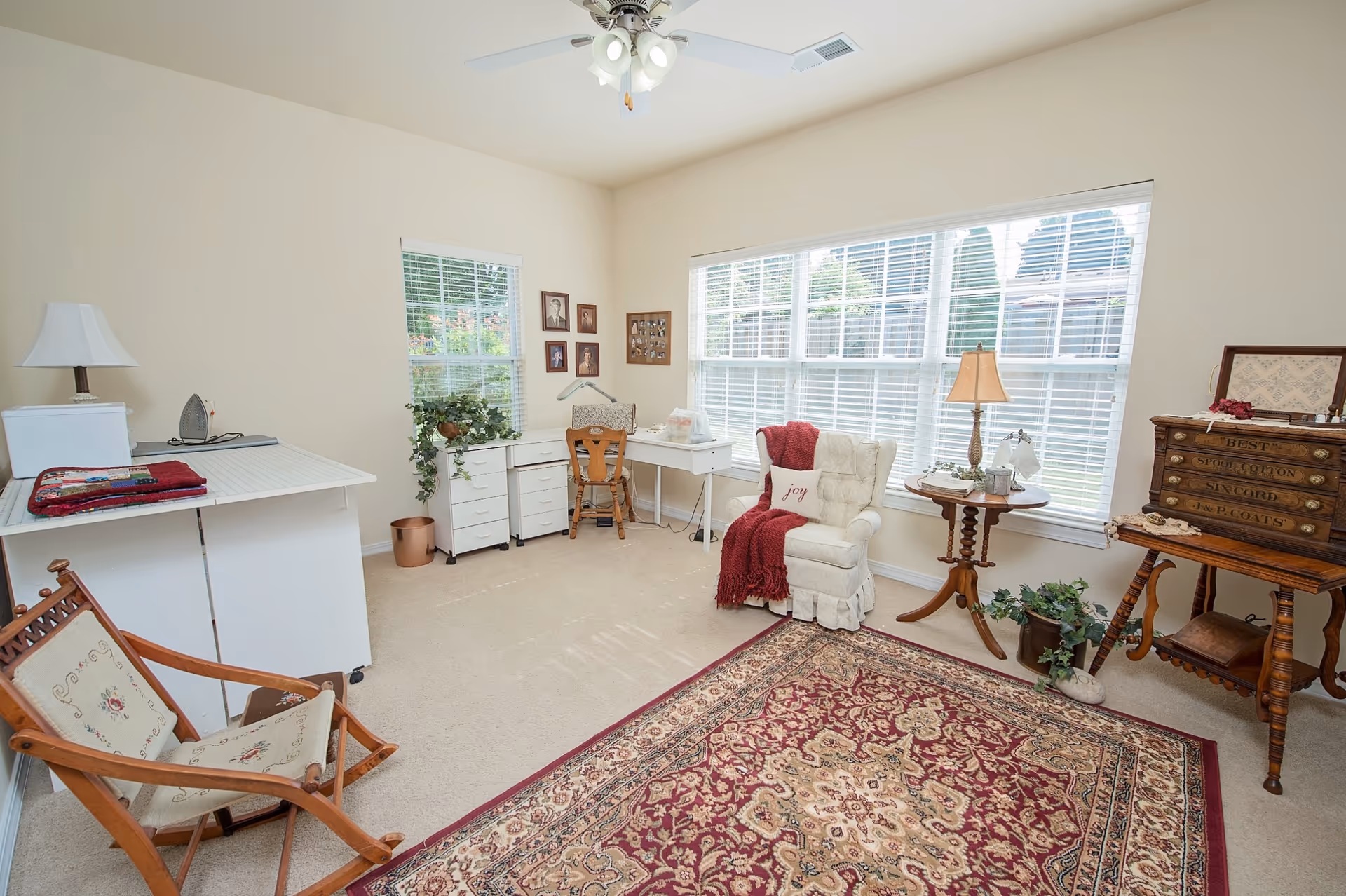 Bright sitting room with a large window, upholstered armchair with a red throw, desk and wooden furniture over a patterned area rug.