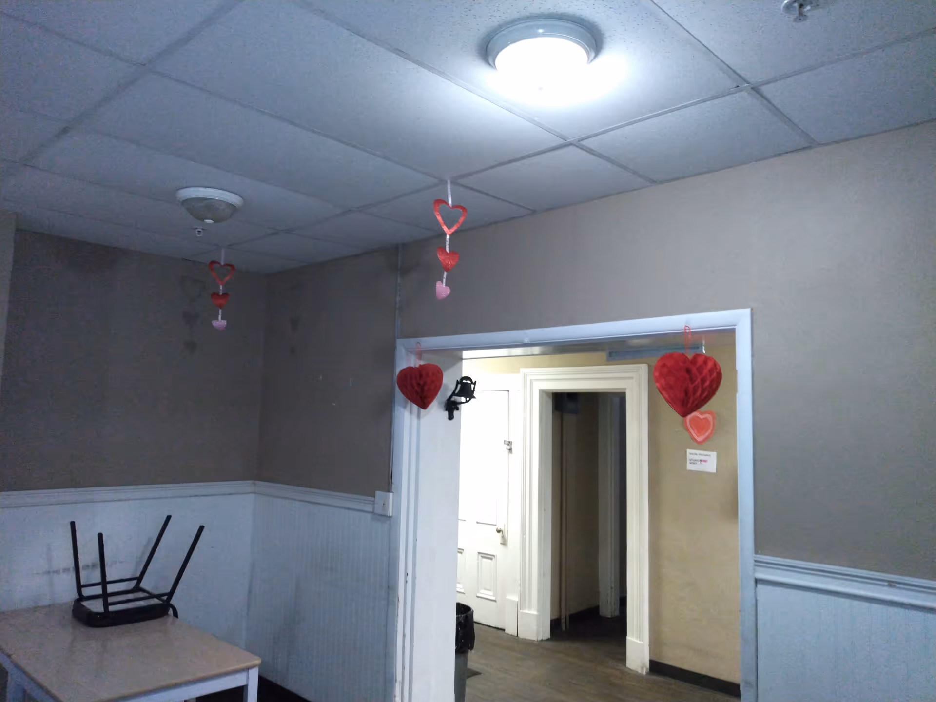 An interior room with beige walls and white wainscoting. A small table with an upside-down chair on top is in the corner. The ceiling has two light fixtures, and red and pink heart-shaped decorations hang from the ceiling and doorway. A hallway with white doors is visible through the doorway.