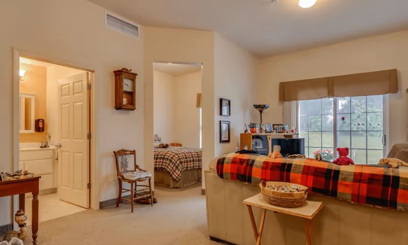 Interior view of a senior living facility showing a cozy living room with a beige couch covered by a red and black plaid blanket, a small wooden table with a basket, a TV stand with framed photos, and a large window with a brown valance. Adjacent to the living room is a bedroom with a bed covered in a plaid blanket, and a bathroom with a sink and mirror visible through an open door. The walls are light-colored, and there is a wooden chair and a wall clock in the hallway area.