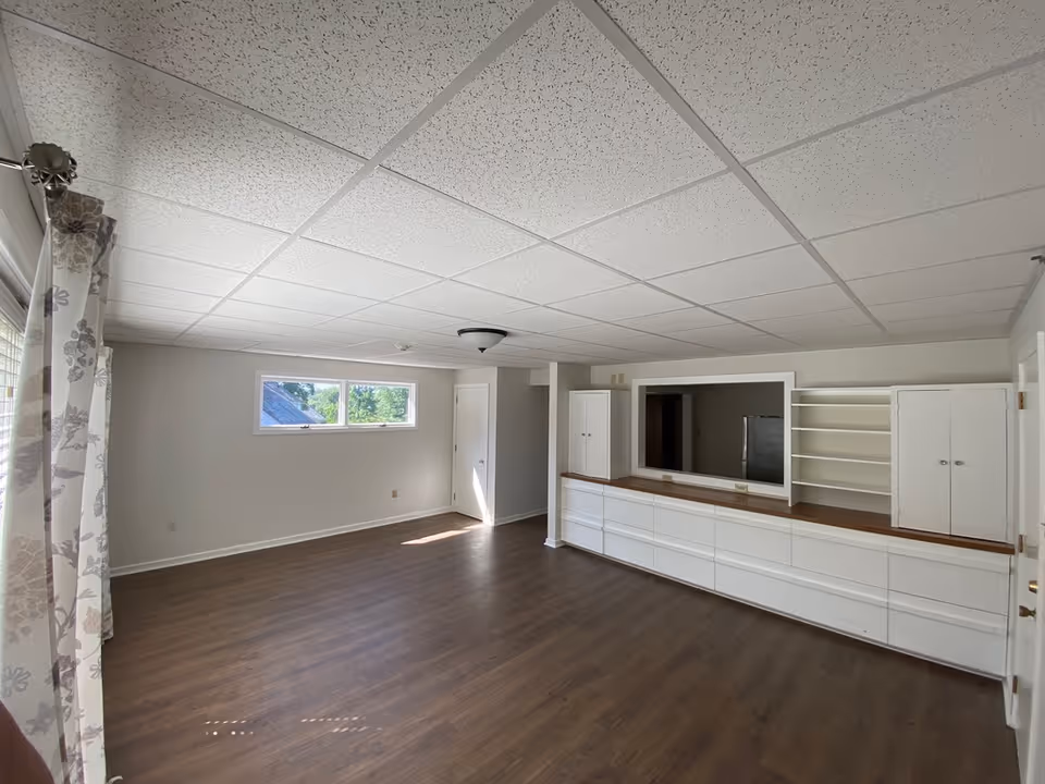 Empty room with wood flooring, white walls, and a drop ceiling with white tiles. There is a large built-in white cabinet with multiple drawers and shelves along one wall, and a small window letting in natural light. Floral curtains hang on the left side near a larger window or door.