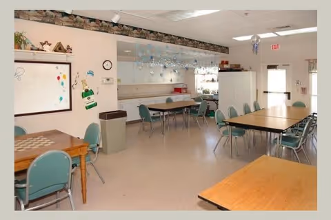 A bright room with several tables and green chairs arranged around them, a whiteboard on the wall with colorful drawings, a clock, and a large mirror reflecting the room. The room has a clean, simple design with overhead fluorescent lighting and a door leading outside.