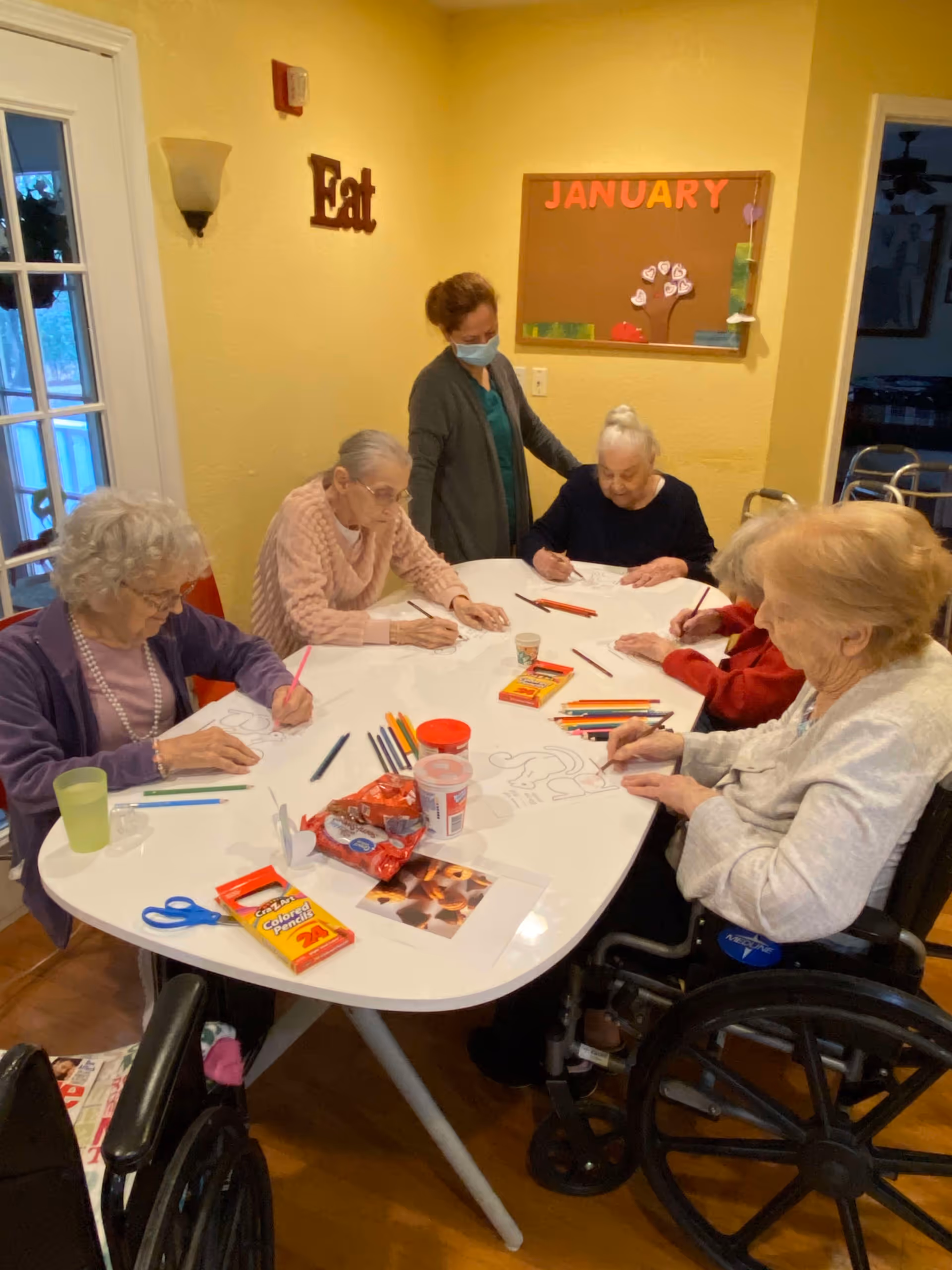 A group of elderly women sitting around a white table engaged in coloring activities with colored pencils and paper. A caregiver wearing a mask stands behind them, assisting. The room has yellow walls with a bulletin board labeled 'JANUARY' and a wall decoration that says 'Eat'.