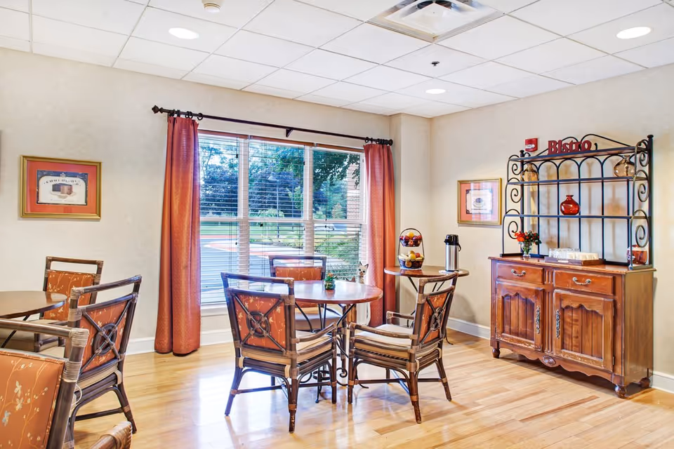 A cozy dining area with wooden tables and chairs featuring orange cushions. There is a large window with red curtains letting in natural light and showing a view of greenery outside. On the right side, there is a wooden cabinet with a black metal rack labeled 'Bistro' holding decorative items and a coffee thermos. The walls are light-colored with framed pictures hanging.