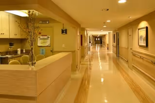 A clean, well-lit hallway inside a senior care facility with beige walls and shiny floors. On the left side, there is a reception desk with a vase of flowers and some chairs. The hallway extends into the distance with handrails on both sides and framed pictures on the walls.