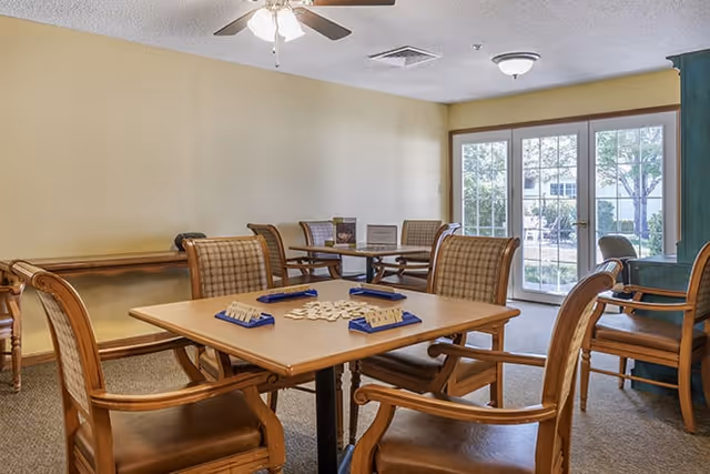 Community activity room with wooden tables and upholstered chairs arranged for a tabletop game and glass doors leading outside.