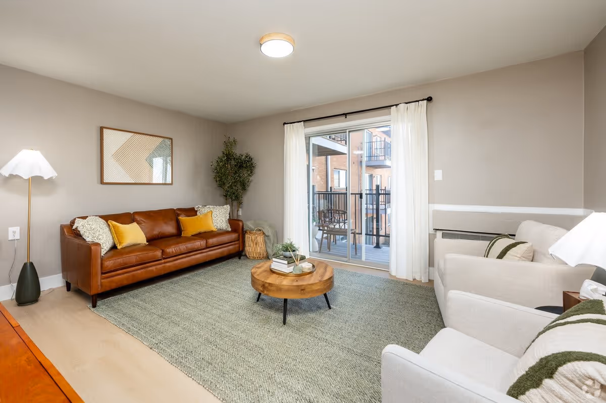 A cozy living room with a brown leather sofa adorned with yellow and patterned pillows, two beige armchairs with green-striped pillows, a round wooden coffee table with books and decor, a floor lamp with a white shade, a large green rug, a potted plant in the corner, and sliding glass doors leading to a balcony with outdoor seating.
