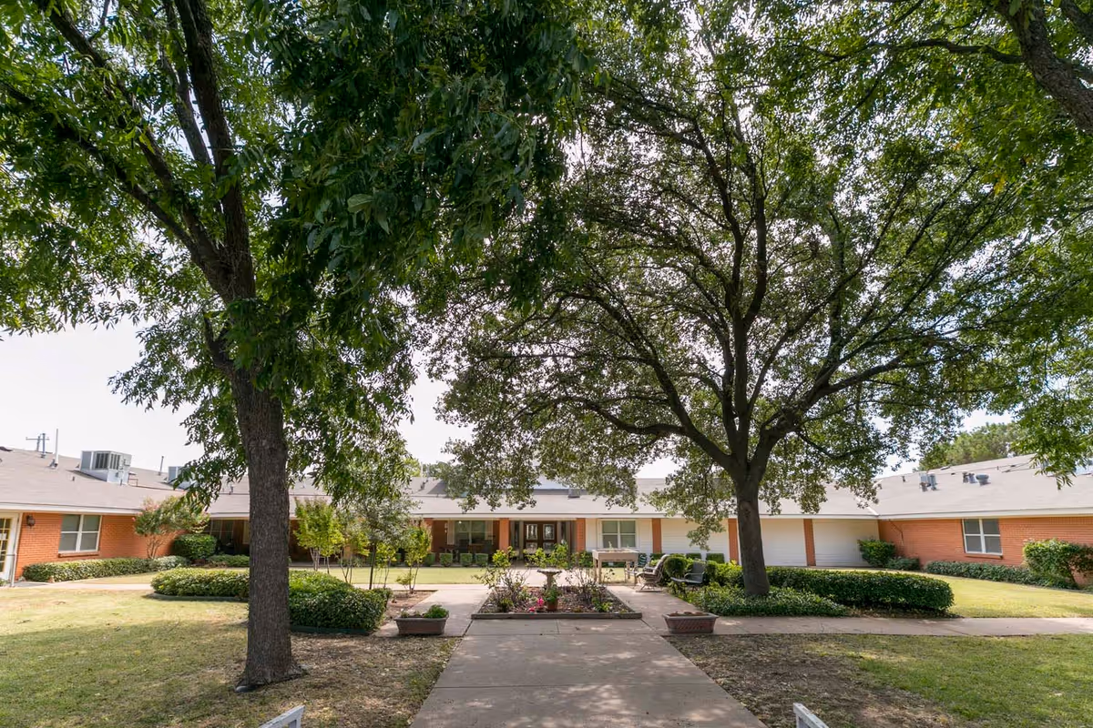 Front courtyard of a single-story brick assisted living building with large trees, a central walkway, planters, and benches.
