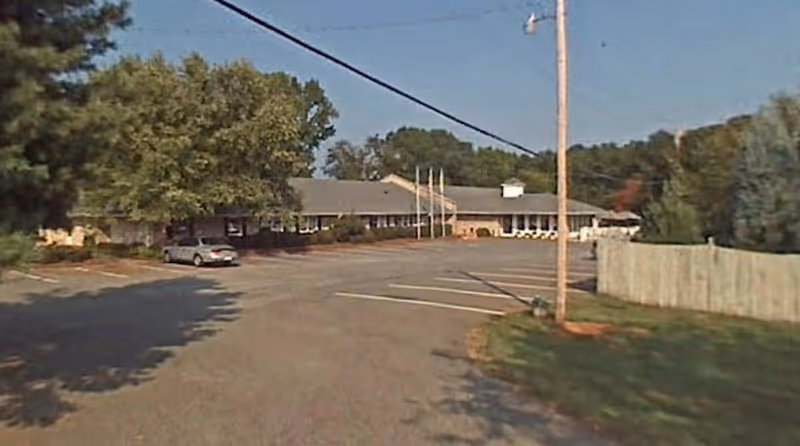 Exterior view of a single-story building with a large parking lot in front. The building is surrounded by trees and a wooden fence on the right side. There is a utility pole with wires running across the image.