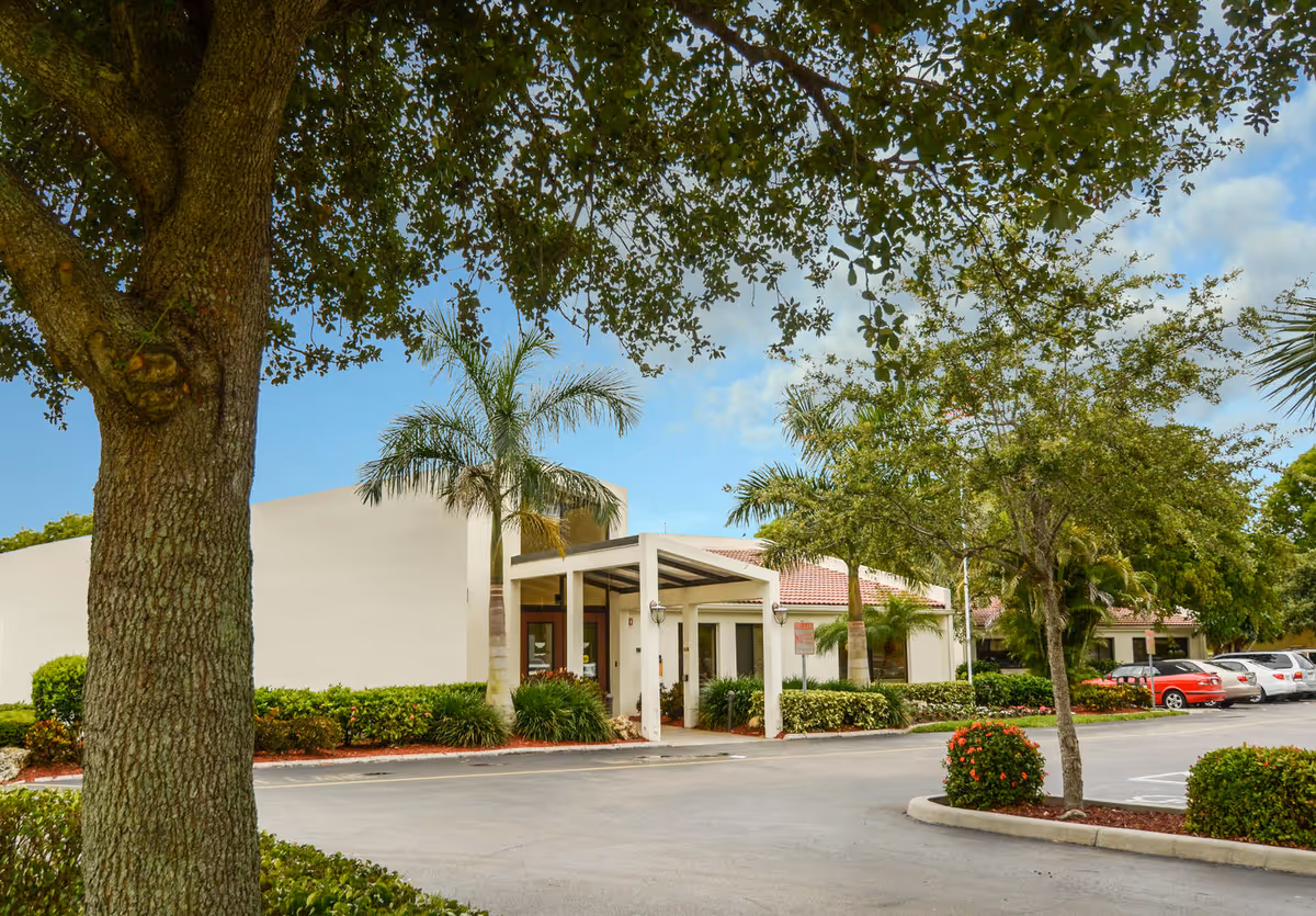 Exterior view of Woodside Health & Rehabilitation Center showing the entrance with a covered drop-off area, surrounded by palm trees, bushes, and a parking lot with several cars. A large tree is in the foreground.