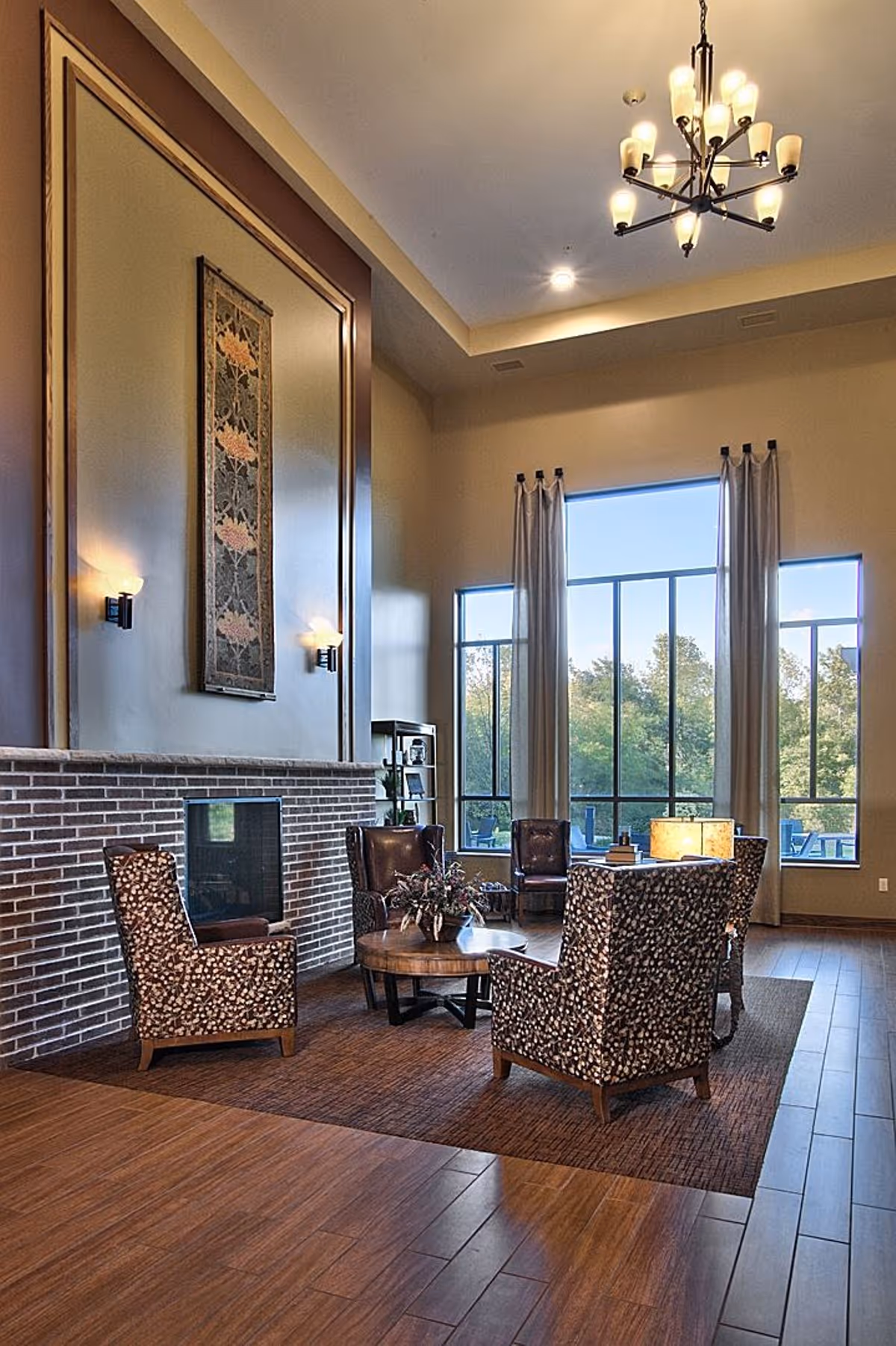 Spacious sitting area with tall windows and high ceiling, a brick fireplace, chandelier, and patterned armchairs around a round coffee table.