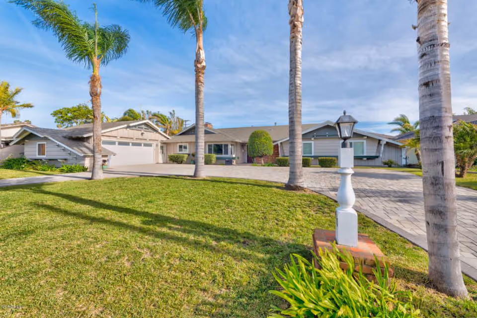 Front exterior view of a single-story residential building with a paved driveway, green lawn, palm trees, and a white lamp post in the foreground under a partly cloudy sky.