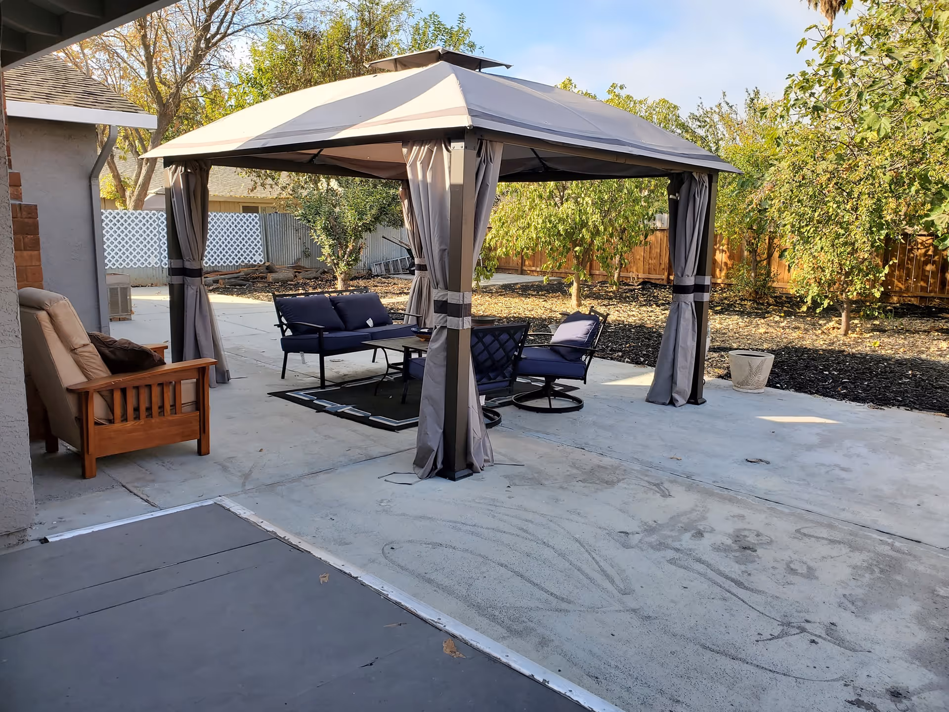 Outdoor patio area with a gray canopy gazebo covering a seating arrangement consisting of a loveseat, two chairs, and a coffee table. There is also a wooden armchair with cushions to the left. The patio is surrounded by trees and a wooden fence in the background.