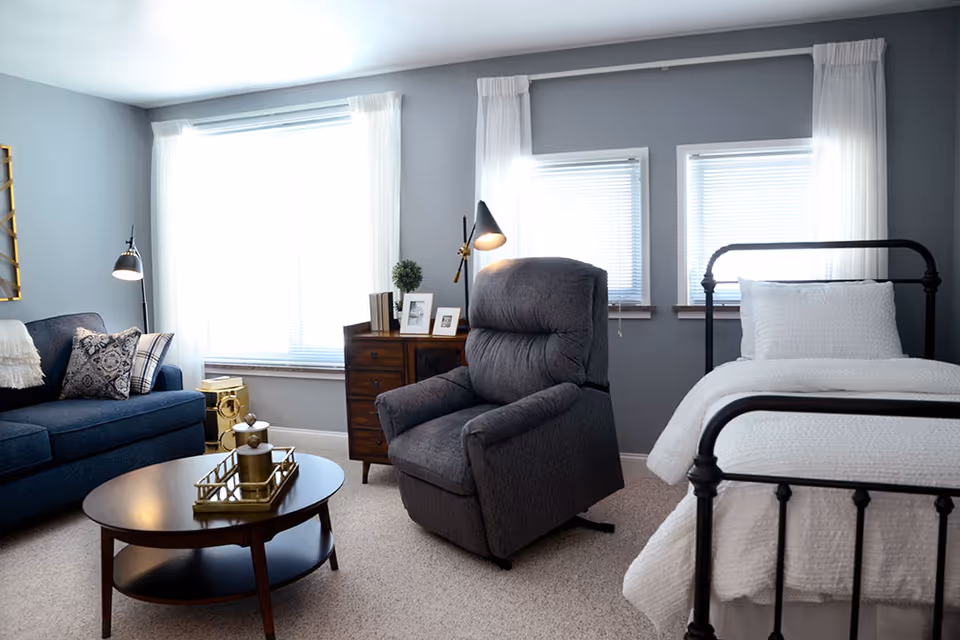 A cozy senior living room and bedroom area with a dark blue sofa adorned with patterned pillows, a round wooden coffee table with decorative items, a gray recliner chair, a wooden dresser with framed photos and a small plant, and a single bed with white bedding and a black metal frame. The room has gray walls and three windows with white curtains allowing natural light to fill the space.