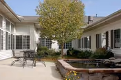 Courtyard patio with a metal table and chairs, a central tree, and a raised brick water feature surrounded by building windows.
