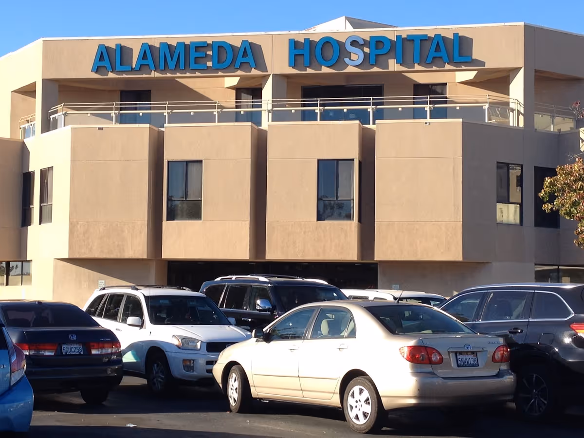Front exterior of Alameda Hospital with large blue signage on a beige facade and several parked cars in the lot.