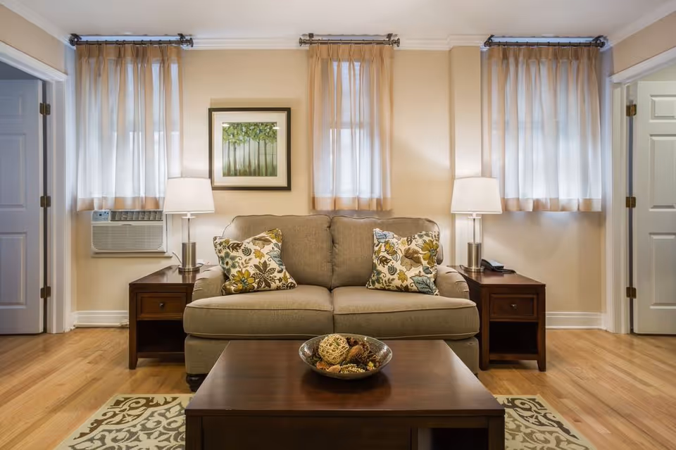 A cozy living room with a beige sofa adorned with two floral-patterned cushions. On either side of the sofa are wooden side tables, each with a modern lamp. Behind the sofa are three windows with sheer curtains, and a framed artwork depicting trees hangs on the wall. A wooden coffee table with a decorative bowl sits in front of the sofa on a patterned rug. The room has light-colored walls and hardwood flooring.
