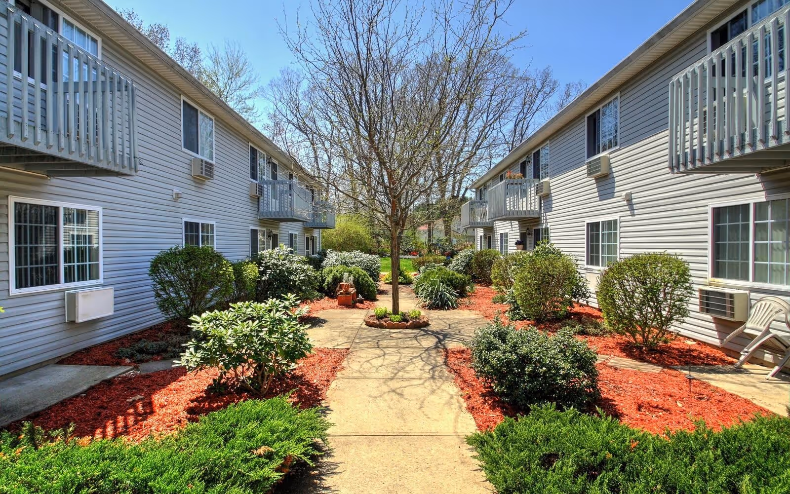 Outdoor courtyard area between two residential buildings with balconies, air conditioning units, and a central walkway lined with bushes, mulch, and a tree in the middle.