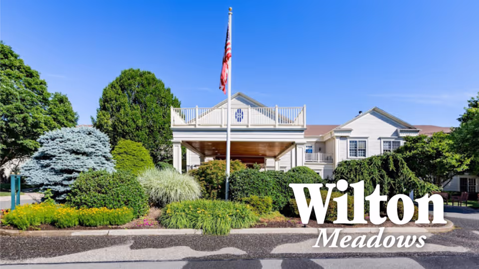Front exterior view of Wilton Meadows Nursing & Rehabilitation facility with well-maintained landscaping, an American flag on a pole, and a clear blue sky.