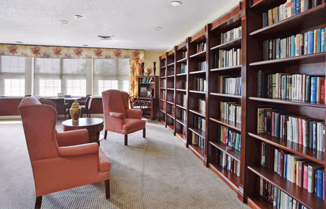 A cozy library room in a senior living facility with large wooden bookshelves filled with books along one wall. Two upholstered armchairs and a round wooden table with a decorative vase are placed near the bookshelves. Large windows with floral curtains allow natural light to fill the room.