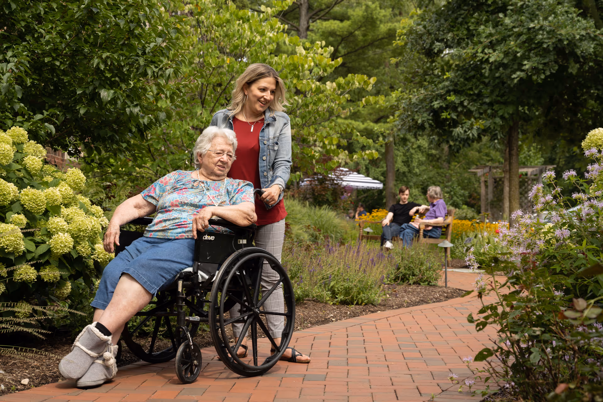An elderly woman in a wheelchair is being pushed by a younger woman along a brick pathway in a lush garden with green trees and flowering plants. In the background, two people are sitting and talking on a bench surrounded by more greenery and flowers.