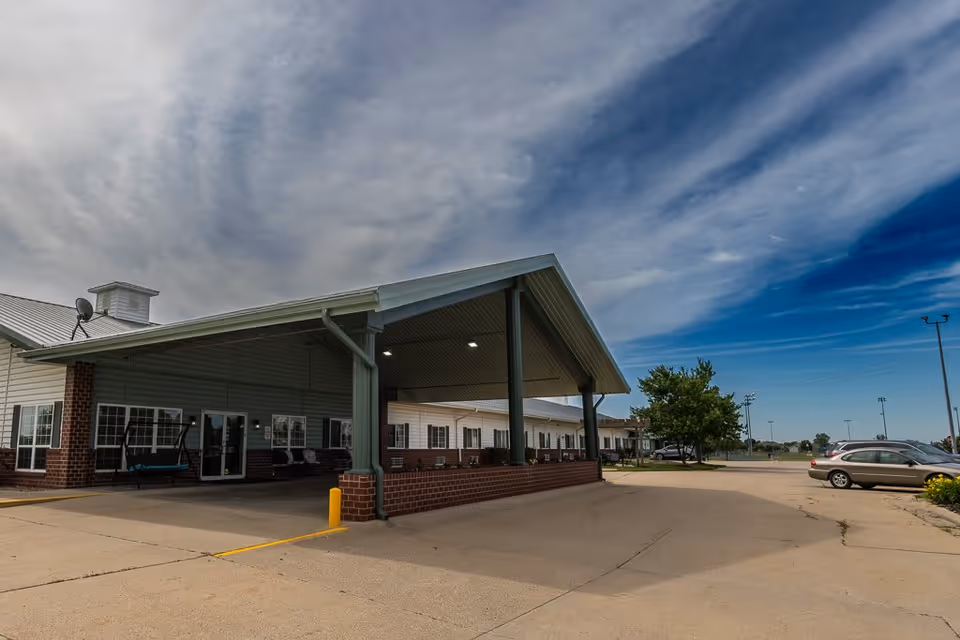 Exterior view of Bailey Pointe at Plattsmouth showing the building entrance with a covered drop-off area, a parking lot with cars, and a partly cloudy sky.