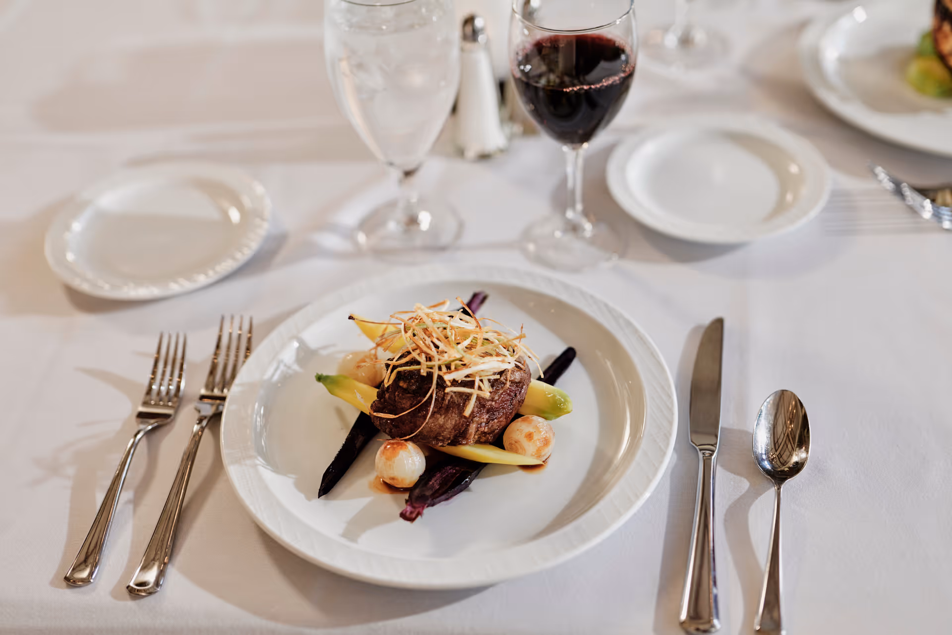 A plated gourmet entrée topped with crispy garnish on a white plate, surrounded by a wine glass, water glass, and silverware on a white tablecloth.