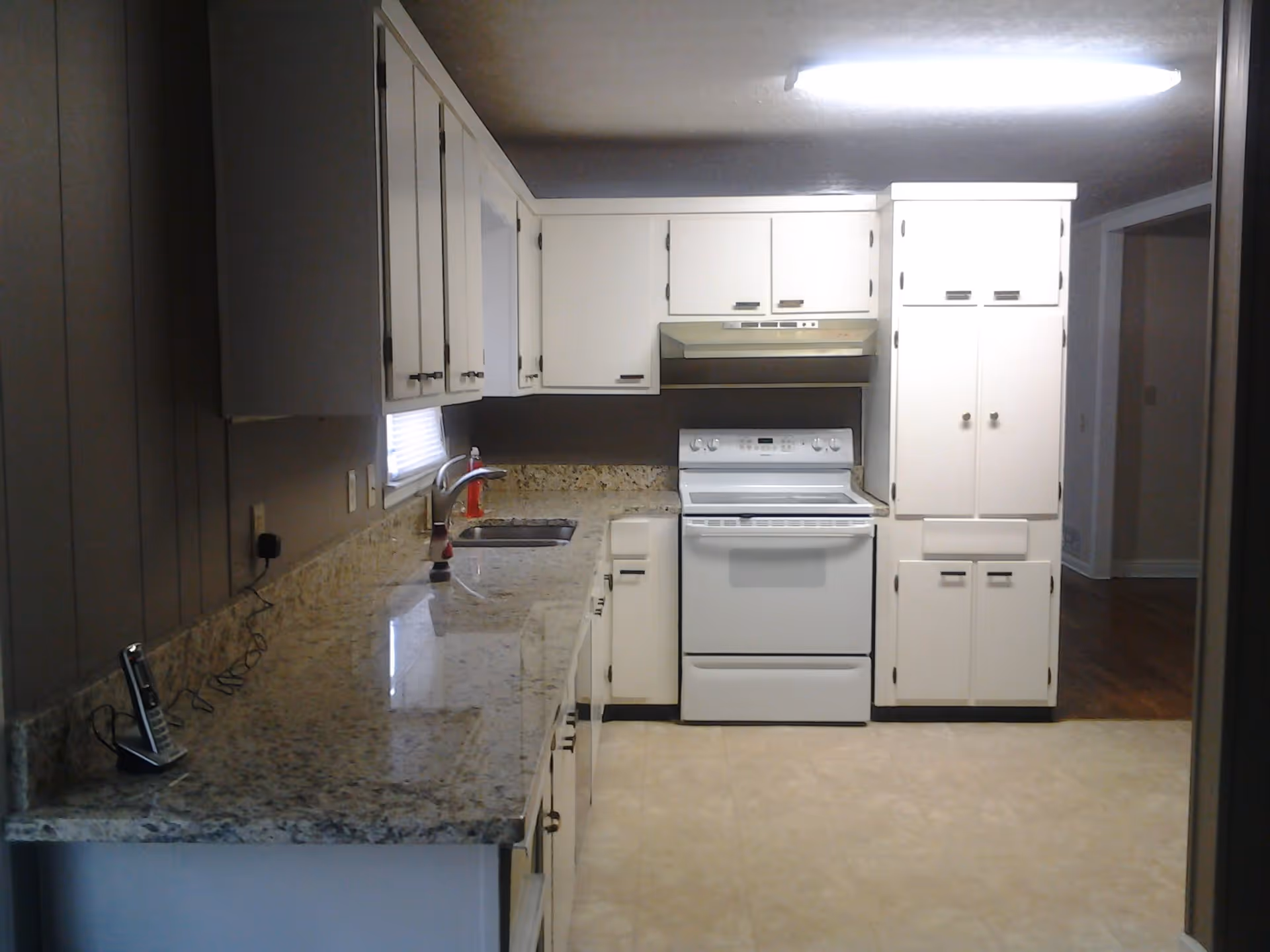 A clean kitchen with white cabinets, a granite countertop, a white electric stove with an overhead range hood, a sink with a faucet, and a cordless phone on the counter. The floor is tiled and there is a fluorescent light fixture on the ceiling.