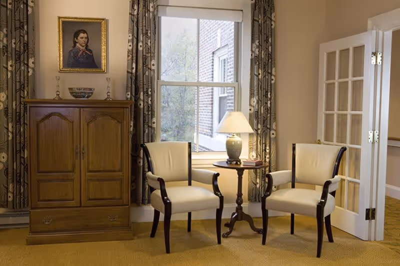 A cozy sitting area with two cream-colored armchairs with dark wooden frames positioned around a small round wooden table holding a lamp and a book. Behind the chairs is a large window with patterned curtains, and to the left is a wooden cabinet topped with decorative items and a framed portrait hanging above it. To the right, a white door with glass panes is partially open, revealing a hallway.