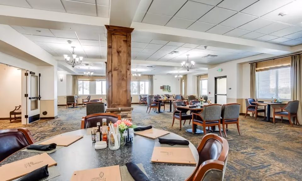 A spacious dining room in Pine Grove Crossing with multiple round and square tables set with menus, black napkins, and condiments. The room features large windows with beige curtains, carpeted floors with a patterned design, and wooden columns. Ceiling lights and chandeliers provide illumination.
