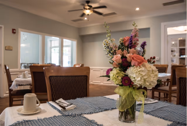 Dining room table set with blue placemats, white dishes and a vase of fresh flowers in a light-filled room with wooden chairs and a ceiling fan.