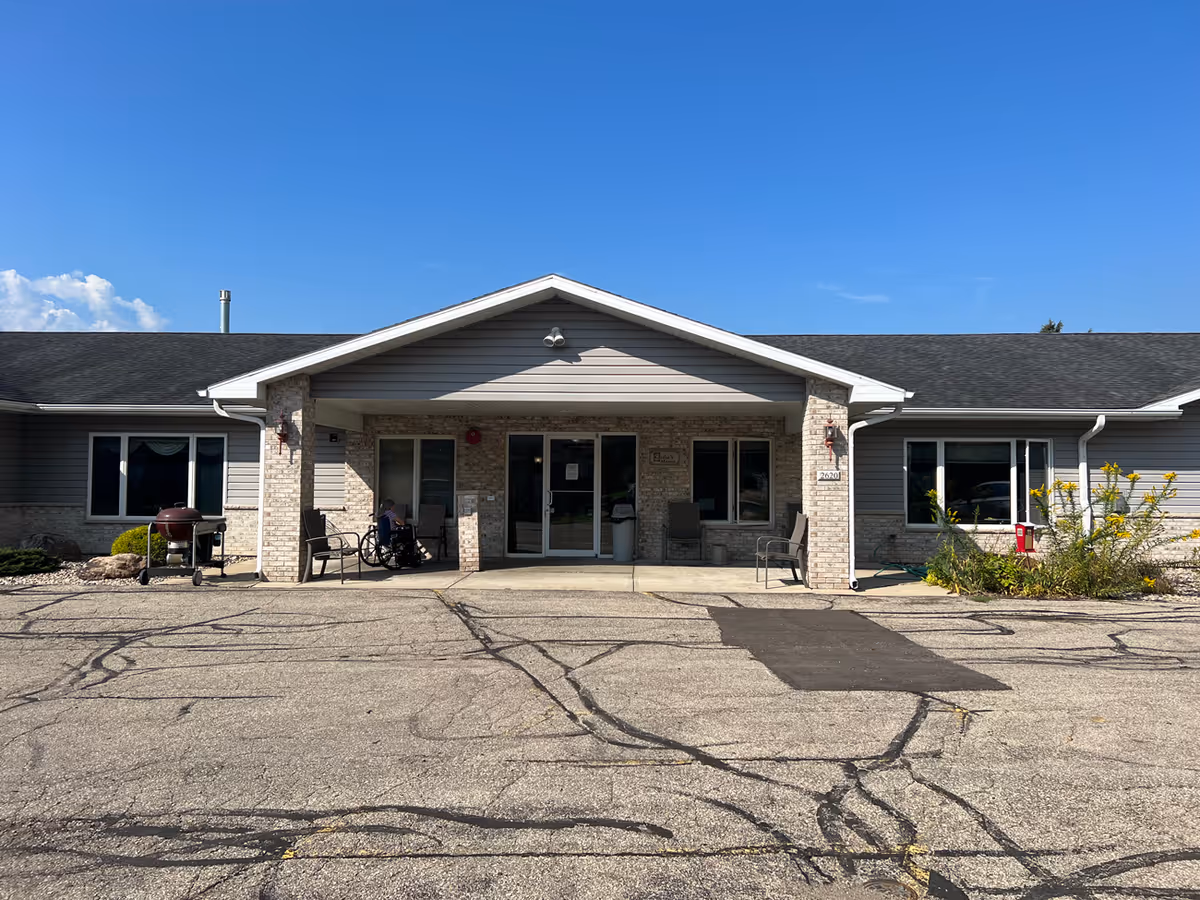 Front entrance of a single-story senior living building with a covered porch, chairs, and a wheelchair visible.