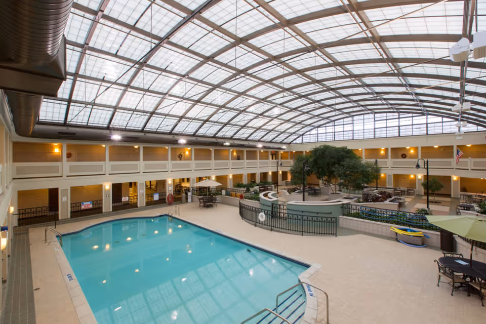 Indoor swimming pool area with a large glass ceiling allowing natural light to fill the space. Surrounding the pool are tables with umbrellas, chairs, and a fenced garden area with trees and plants. The area is enclosed by a two-story structure with doors and windows, and warm lighting fixtures are mounted on the walls.